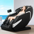 A woman reclines in a black and silver massage chair in a bright indoor setting with ocean scenery visible in the background.
