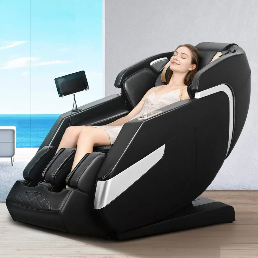 A woman reclines in a black and silver massage chair in a bright indoor setting with ocean scenery visible in the background.